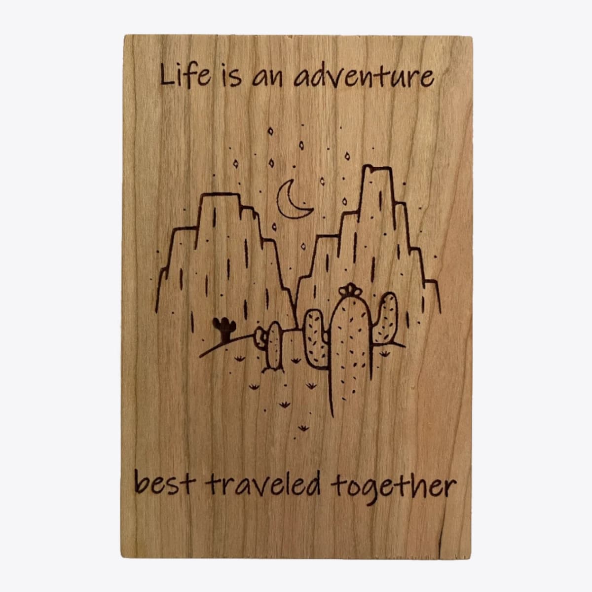 A wooden card that is laser engraved with an image of desert mountains at night and text that says life is an adventure best traveled together.